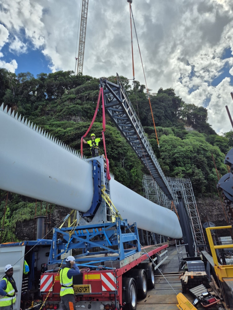A wind turbine blade on an auxiliary structure and a trailer, with workers in high-visibility vests next to it