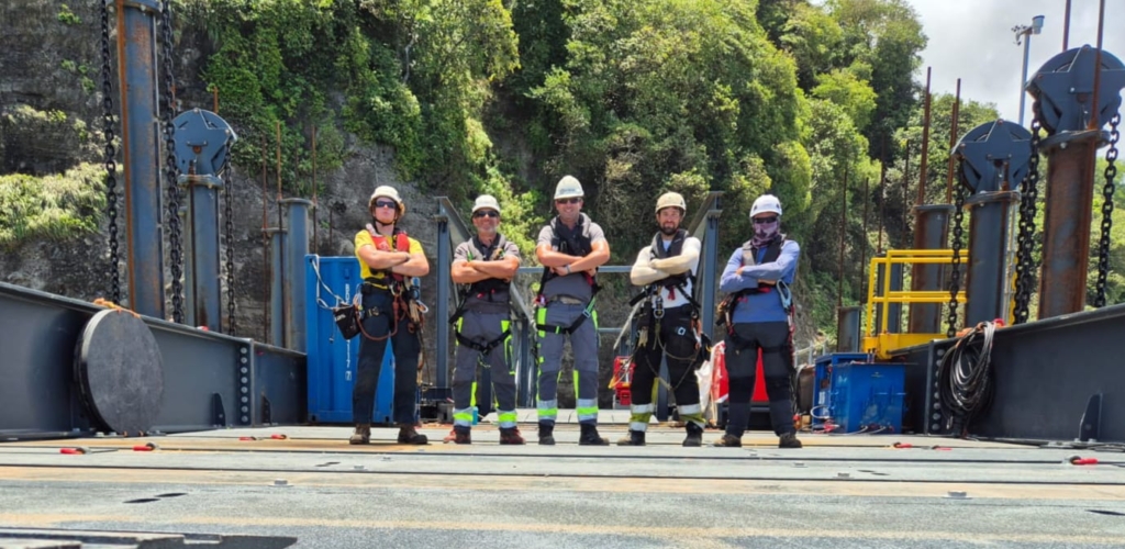 Five workers in safety harnesses on a barge deck with a green hillside behind them