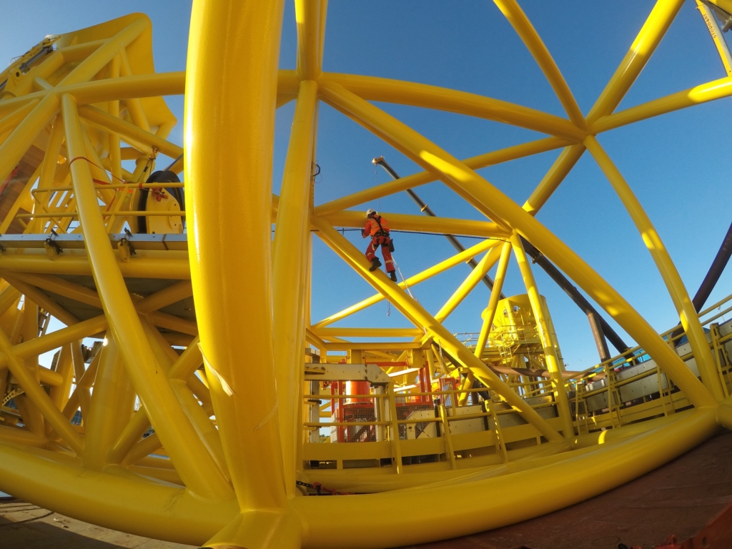 Technicien au milieu du treillis jaune d'une structure offshore, sur un ciel bleu