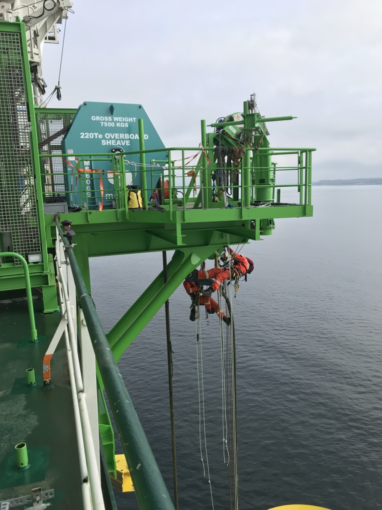 Techniciens sur cordes le long de la coque verte d'un navire au-dessus de l'eau, des personnes au garde-corps sur le pont