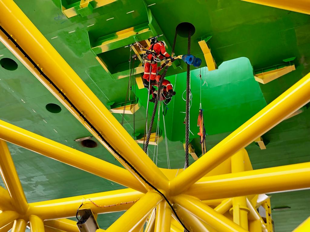 Technicien sur cordes sous une coque verte, parmi les tubes jaunes d'une structure